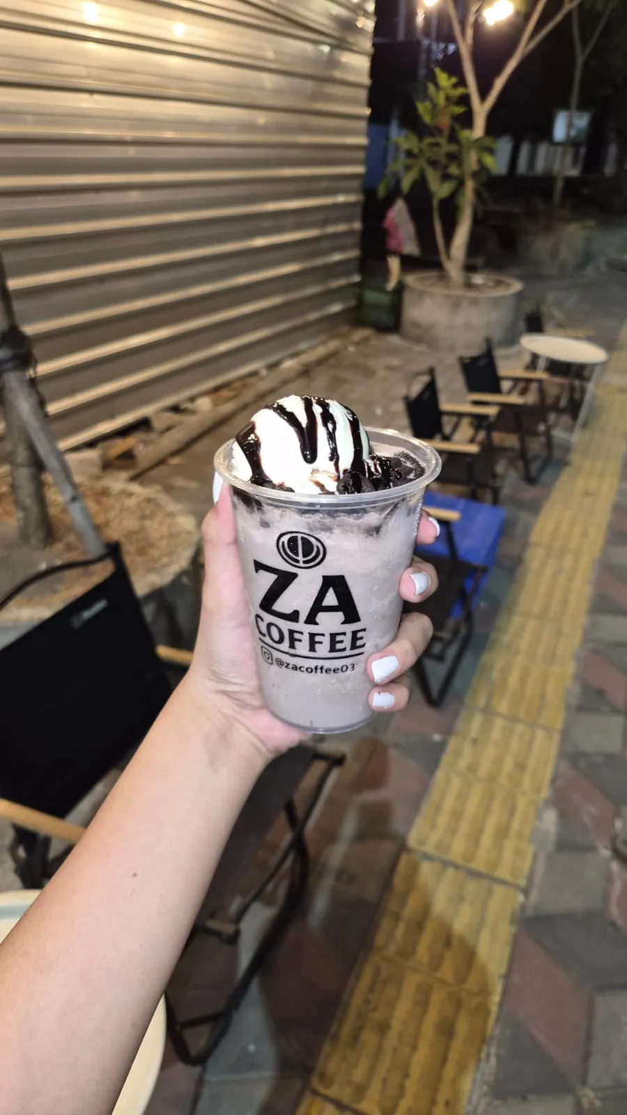 Oreo frappe dipegang di trotoar depan ZA Coffee pada malam hari, kursi lipat camping terlihat di latar belakang