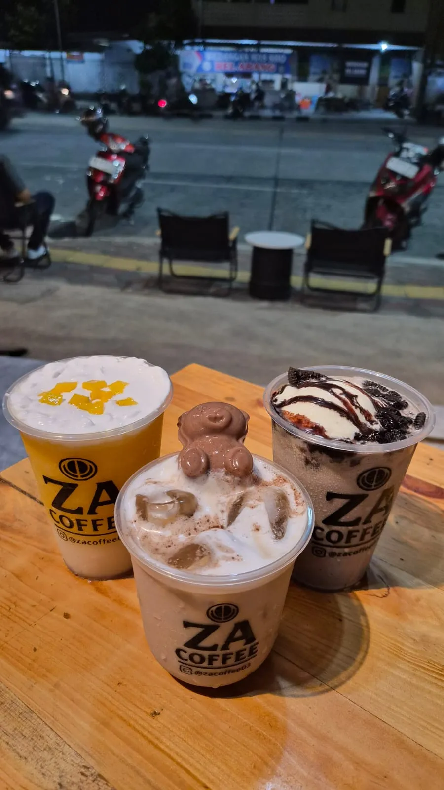 Tiga gelas minuman ZA Coffee bersama di meja kayu, motor dan jalanan malam di latar