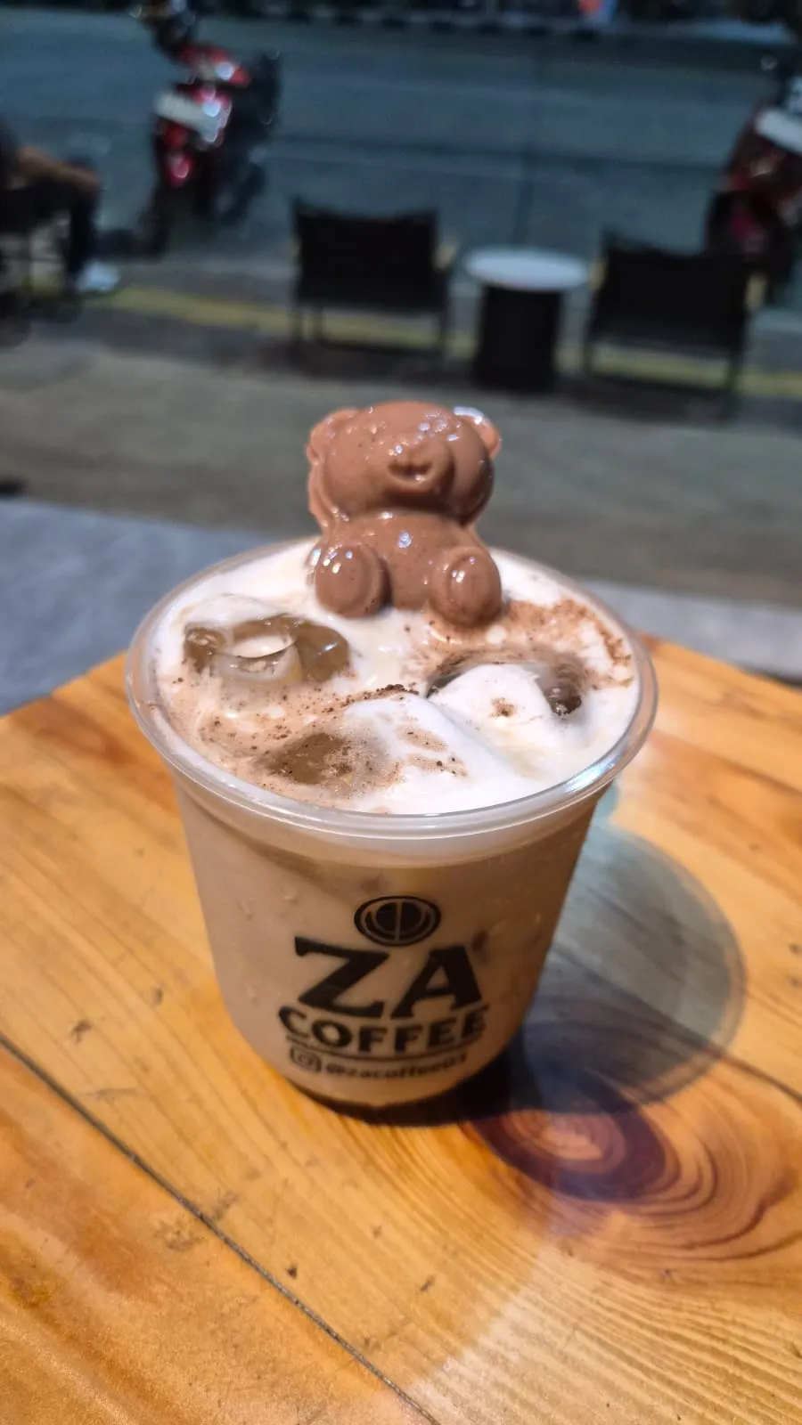 Es kopi ZA Coffee dengan cokelat berbentuk beruang di atas busa, dipegang di meja kayu dengan jalanan malam di latar belakang