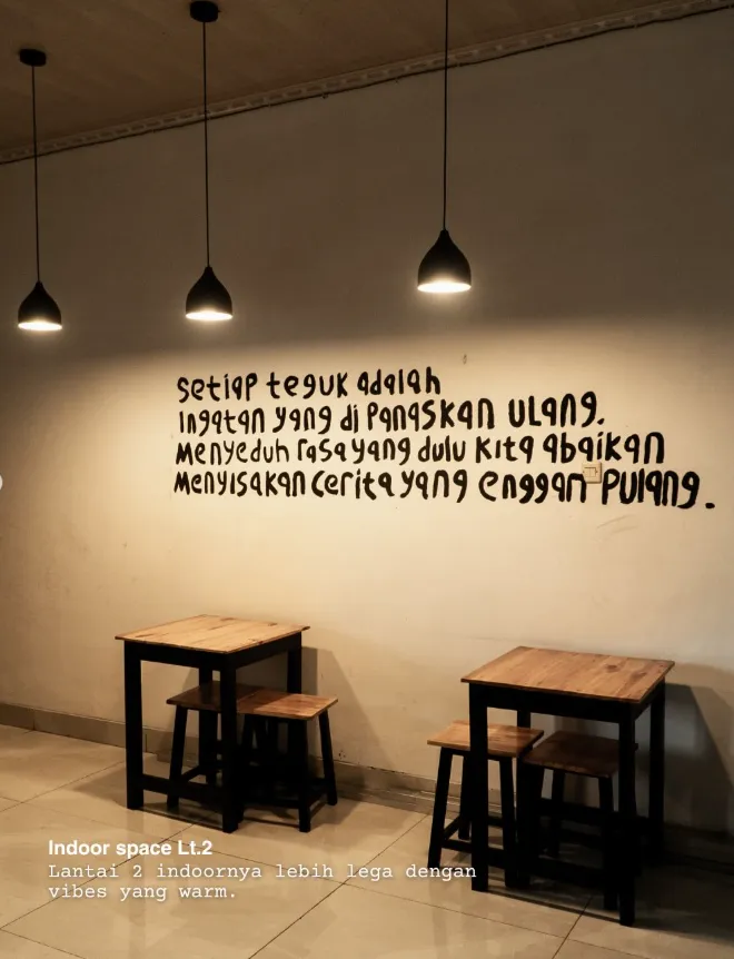 Dinding indoor lantai dua ZA Coffee bermural tulisan tangan: setiap teguk adalah ingatan yang di panaskan ulang. menyeduh rasa yang dulu kita abaikan, menyisakan cerita yang enggan pulang. Tiga lampu gantung dan meja kayu kecil di bawahnya